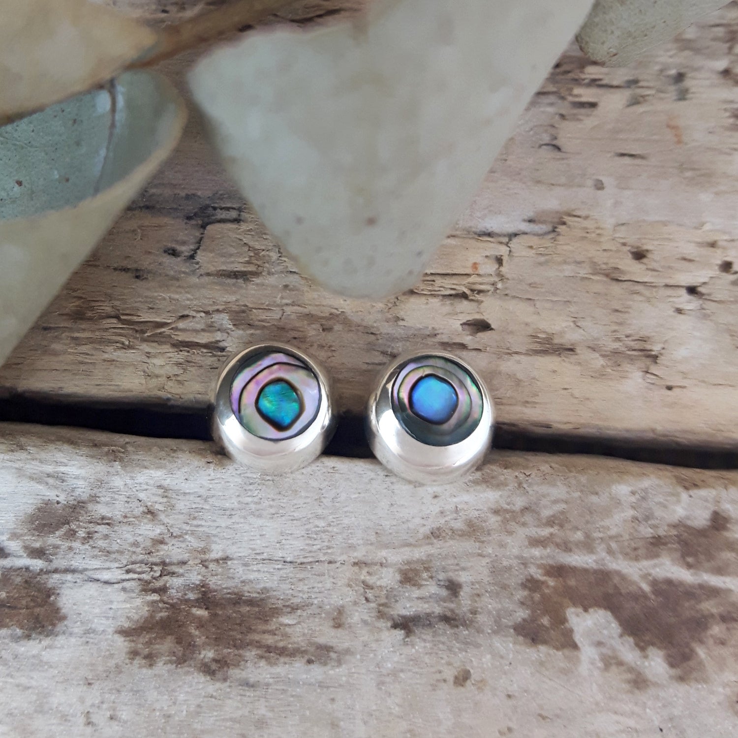 Close-up pair of dainty abalone circle stud earrings in 925 sterling silver, vivid teal and blue iridescent shell, styled on driftwood.