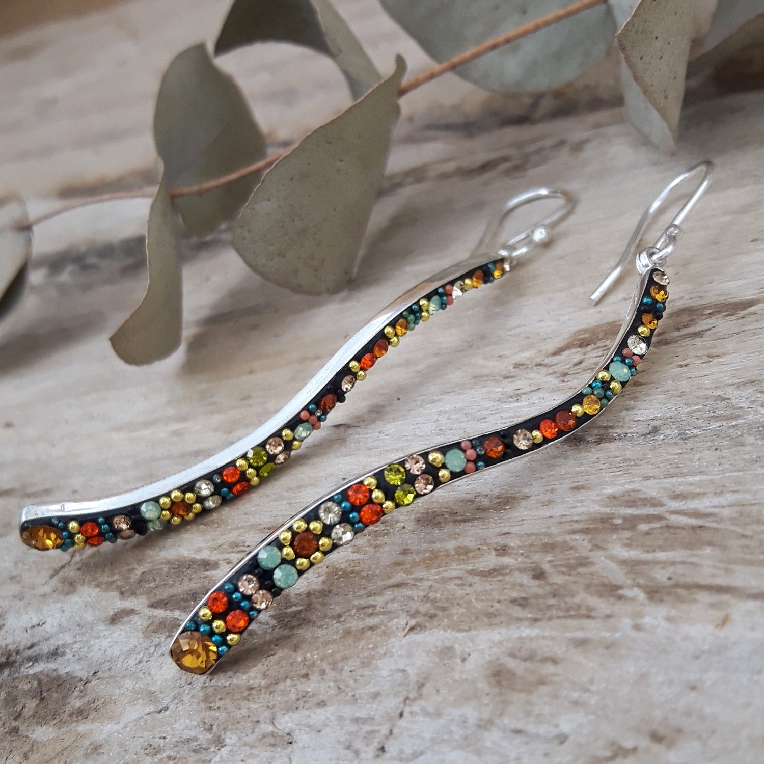 Sterling silver bohemian earrings with multicoloured micro-pearls and crystals on a wooden surface with eucalyptus leaves.