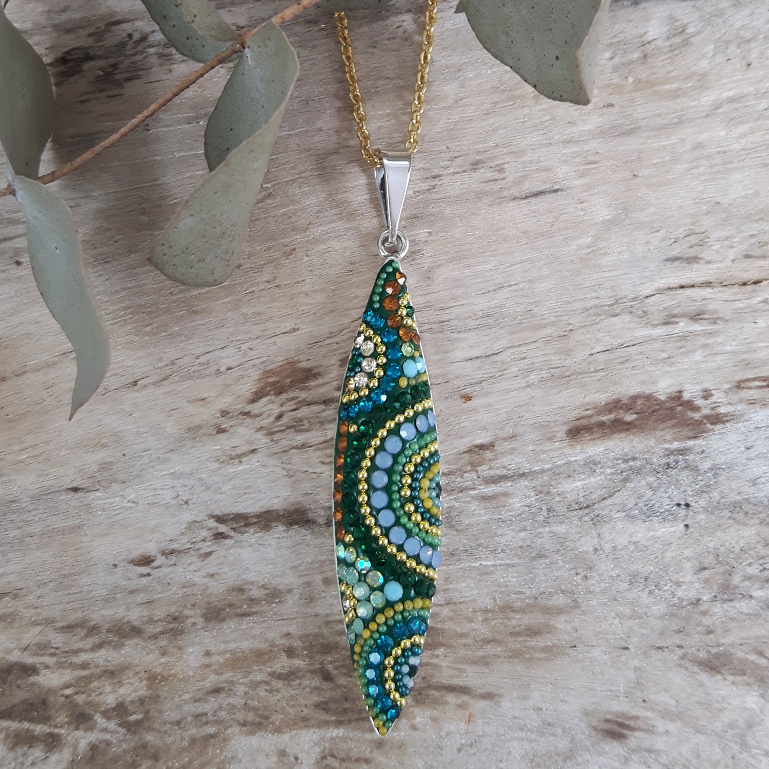 Hand-applied micro-pearl and crystal pendant in green, gold and amber on a gold chain on a wooden surface with leaves