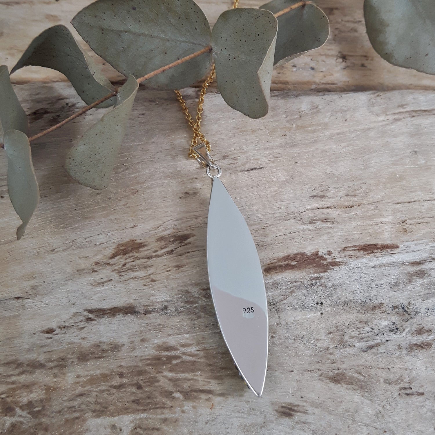 Sterling silver feather-shaped pendant on a gold chain with eucalyptus leaves on a wooden surface