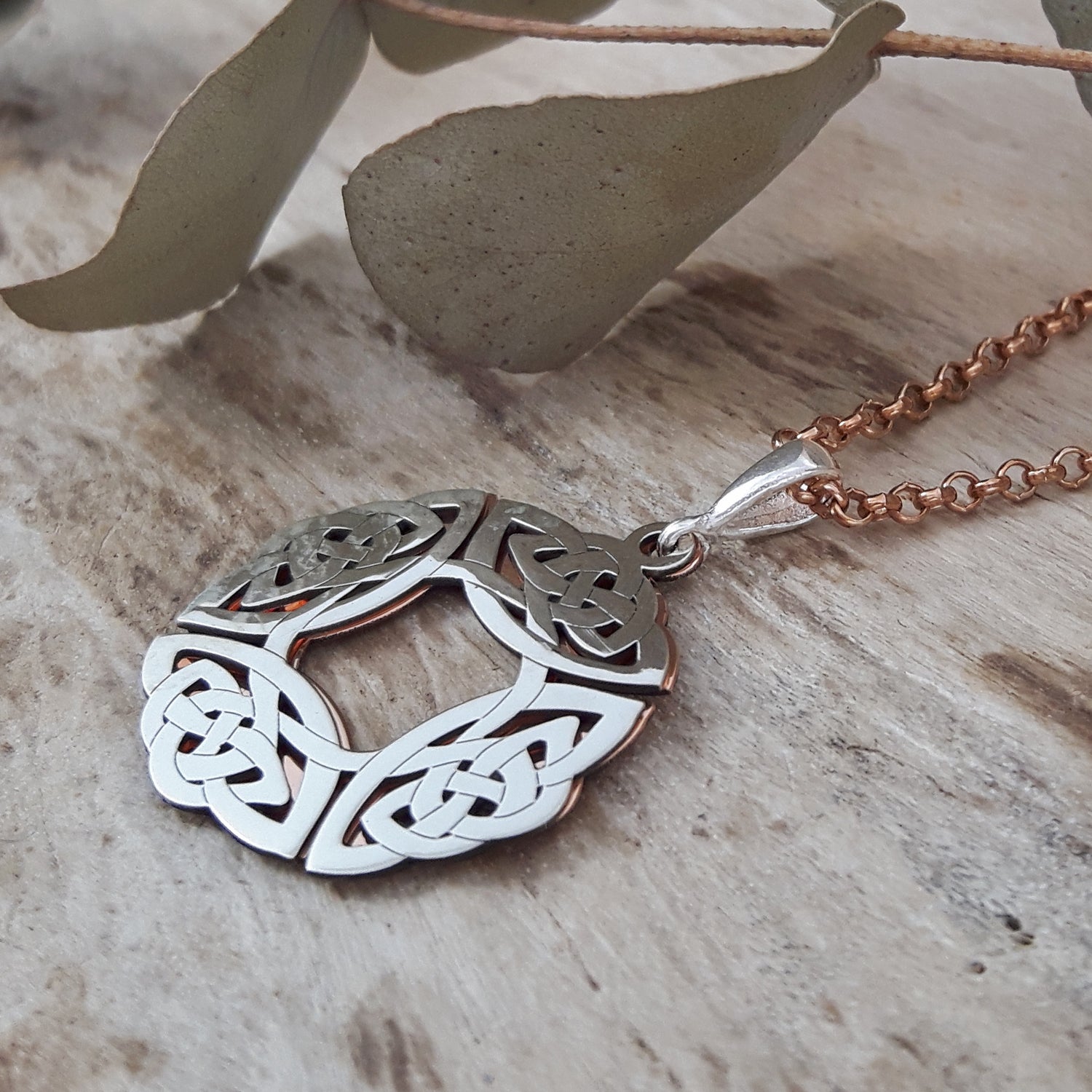 Sterling silver & copper pendant with intricate designs on a wooden surface with leaves.
