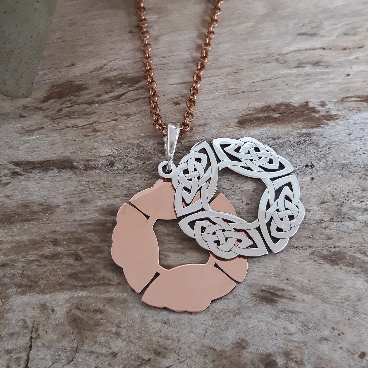 Two parts of a pendant, one copper and one sterling silver, on a rose gold chain against a wooden background.