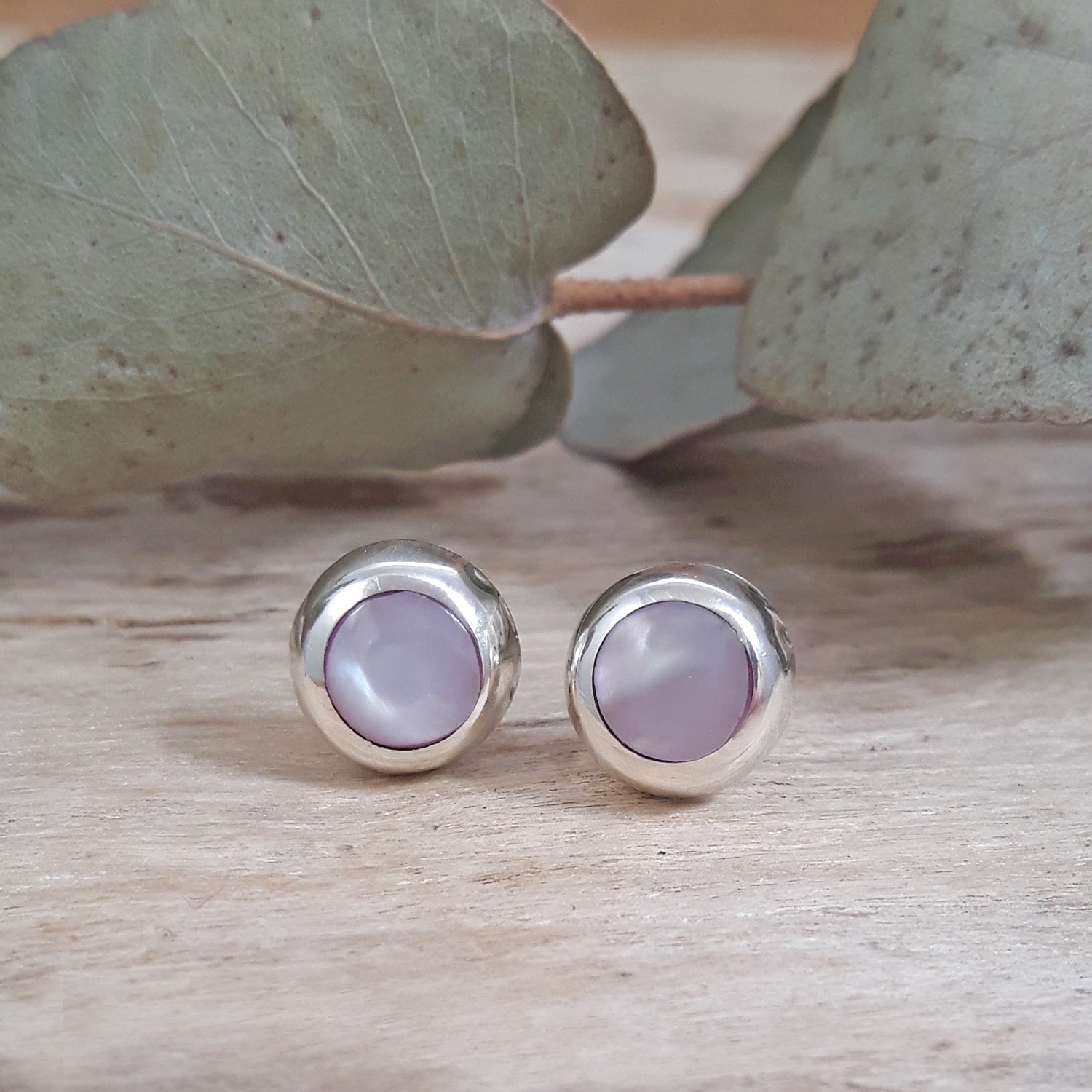 Dainty pink mother of pearl circle stud earrings in 925 sterling silver, soft blush shell, flat-lay on wood with eucalyptus