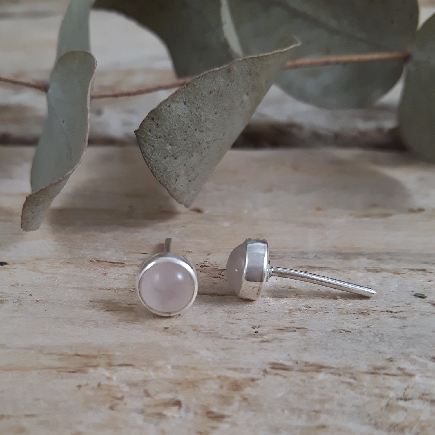 Sterling silver stud earrings with pink Rose Quartz gemstones on a wooden surface with eucalyptus leaves.