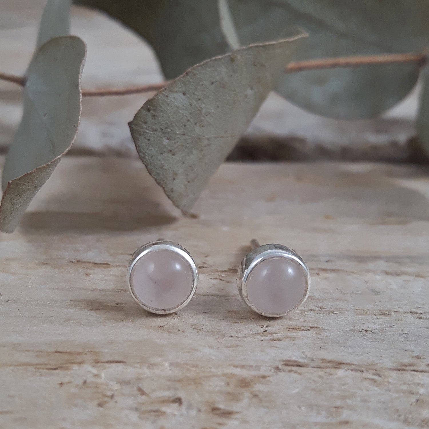 Pair of sterling silver earrings with rose quartz stones on a wooden surface with eucalyptus leaves.