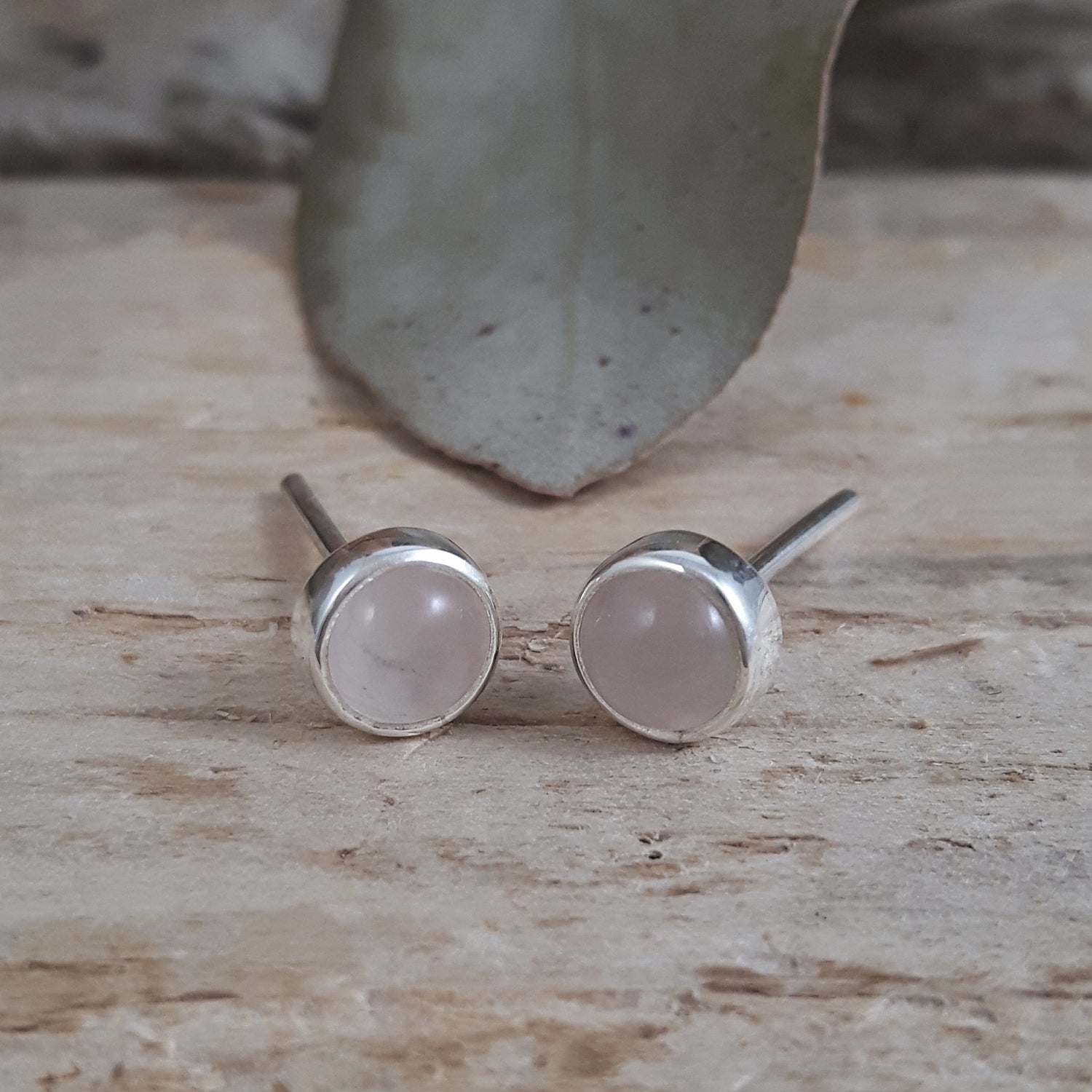 Pair of rose quartz stud earrings on a wooden surface with a stone in the background.