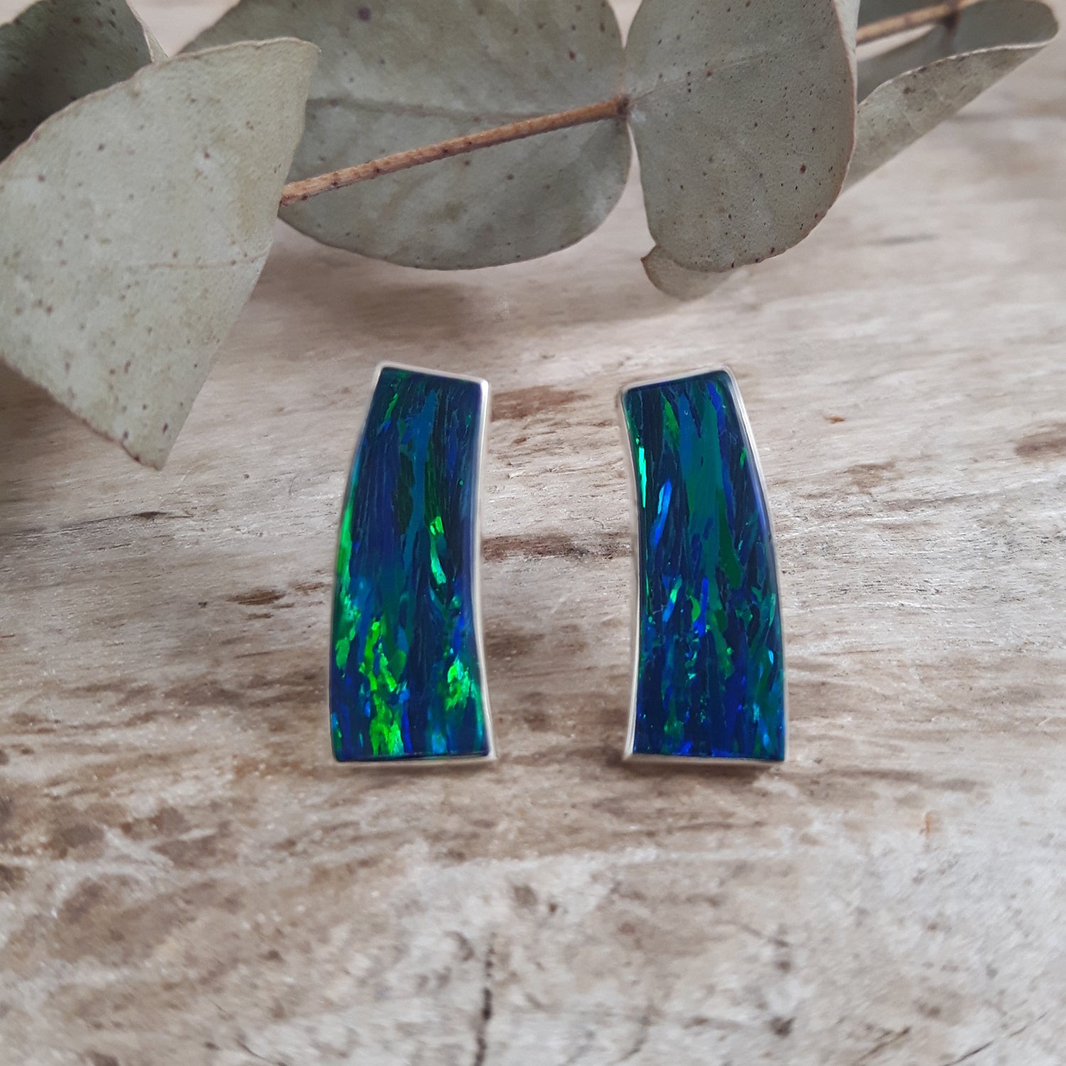 Pair of emerald cultured opal stud earrings on a wooden surface with eucalyptus leaves.