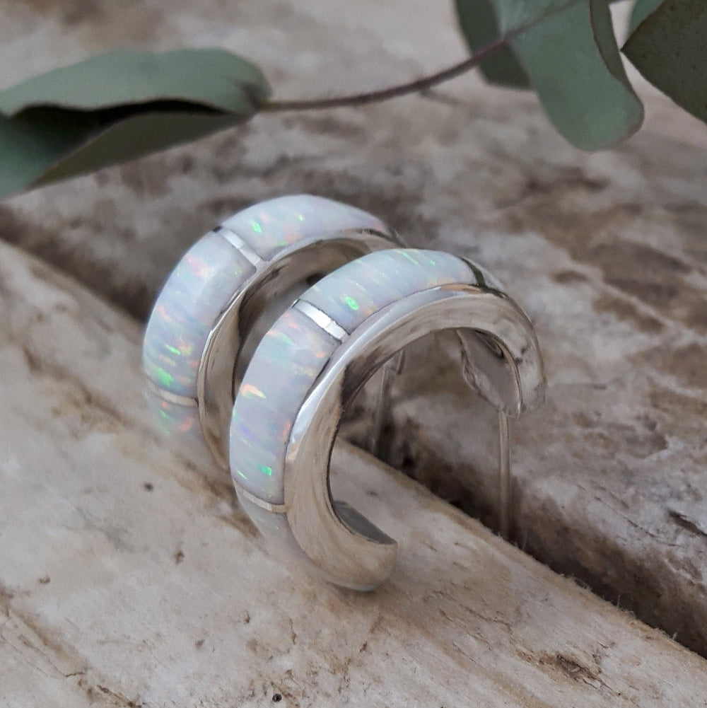 A pair of white hoop earrings with sparkling white cultured opal, set in sterling silver hoops, displayed on a wooden surface with green foliage in the background.