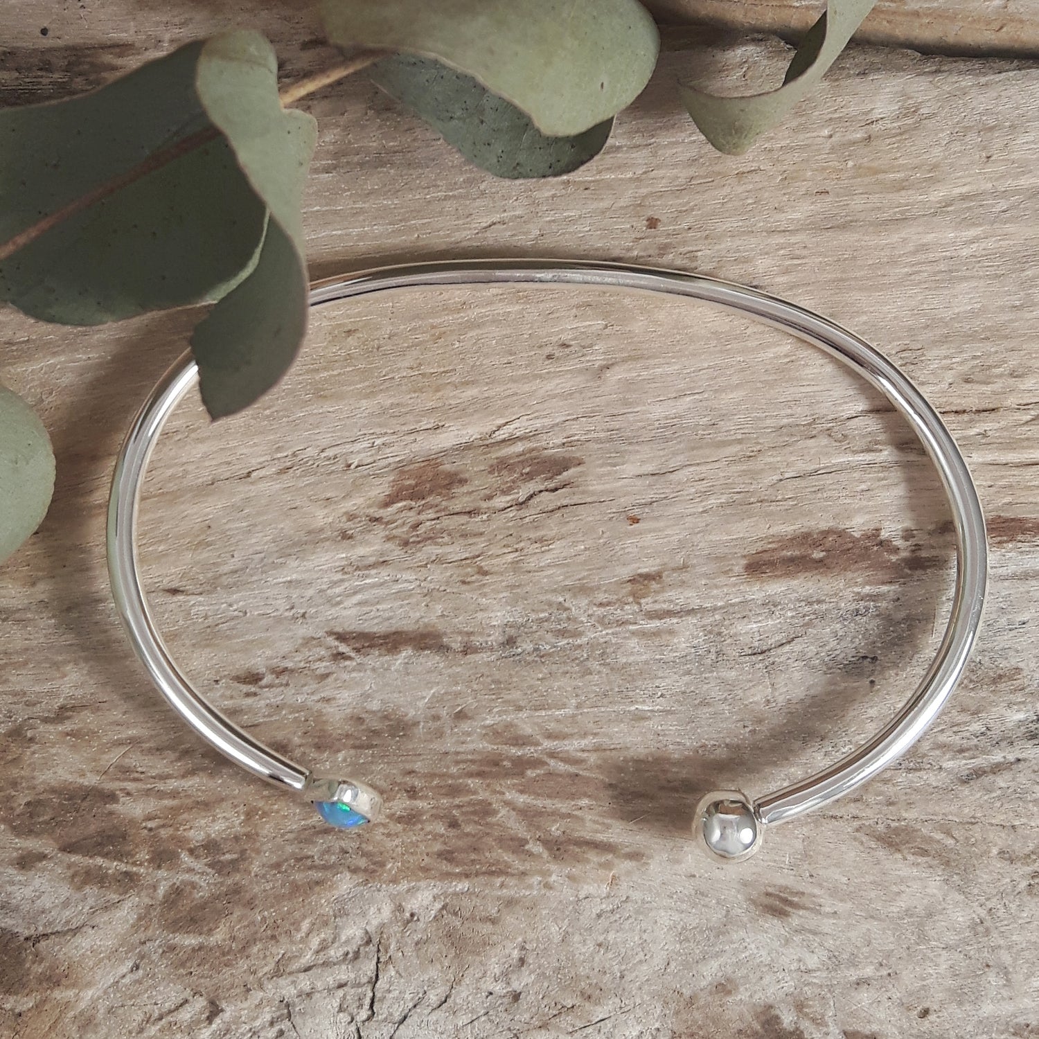 Silver open cuff bracelet with a turquoise gemstone on a wooden surface with eucalyptus leaves.
