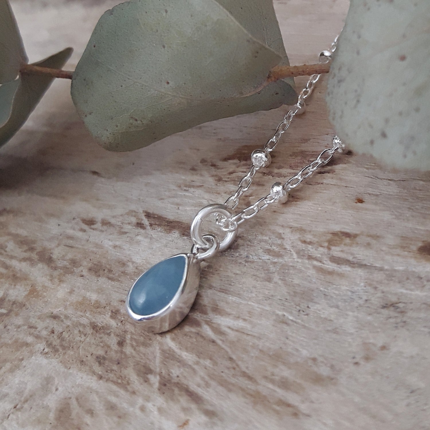 Silver chain necklace with an aquamarine gemstone teardrop pendant on a wooden surface with eucalyptus leaves.