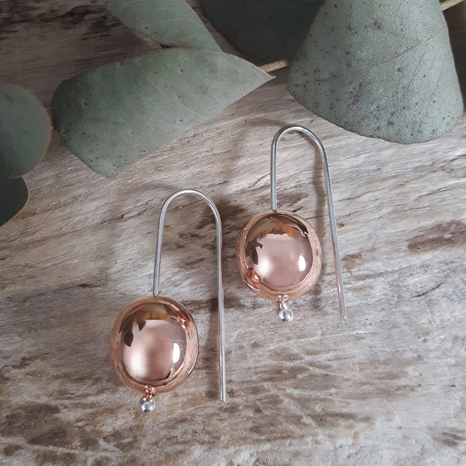 Pair of copper and silver earrings on a wooden surface with green leaves in the background.
