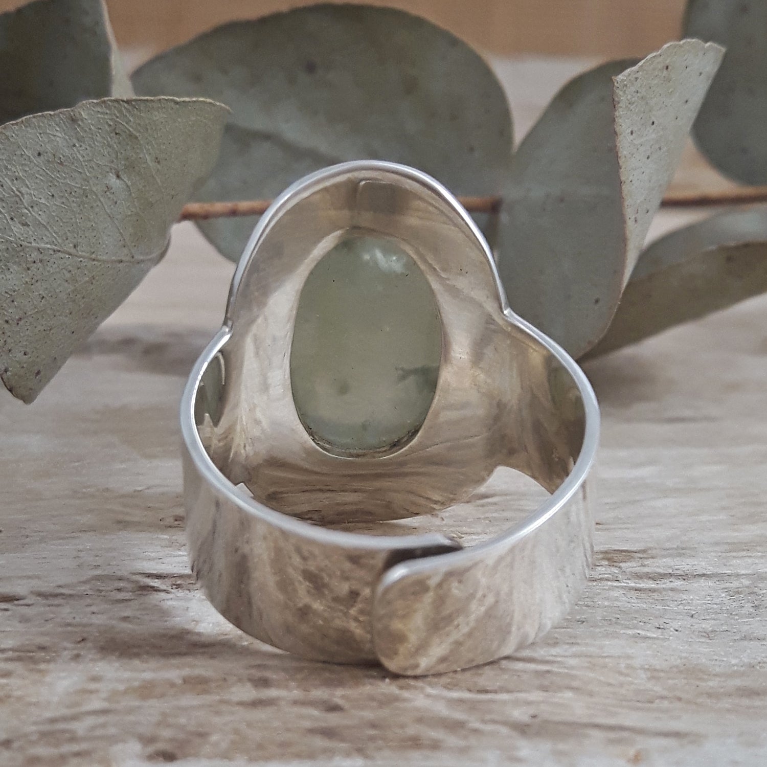Back of a sterling silver ring with a central prehnite stone on a wooden surface with eucalyptus leaves.