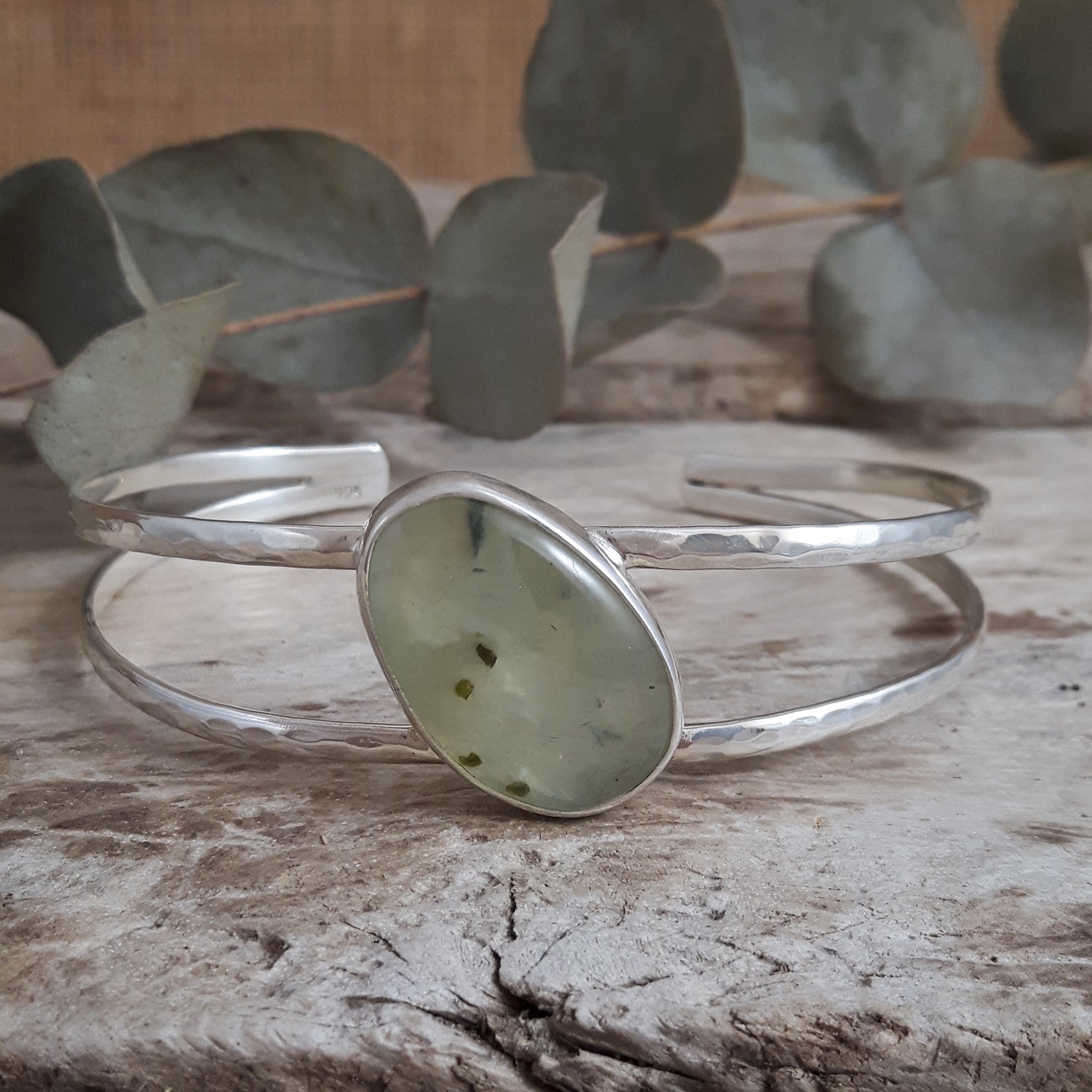 Silver cuff bracelet with a natural prehnite stone focal point on a wooden surface with eucalyptus leaves in the background.