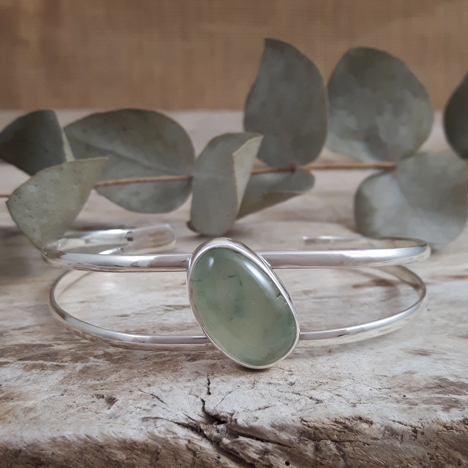 Sterling silver bracelet with a green prehnite stone on a wooden surface with eucalyptus leaves.