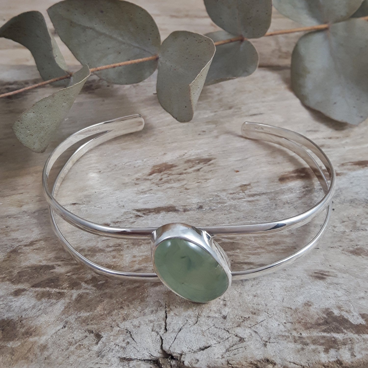 Sterling silver bracelet with a green prehnite stone on a wooden surface with eucalyptus leaves.