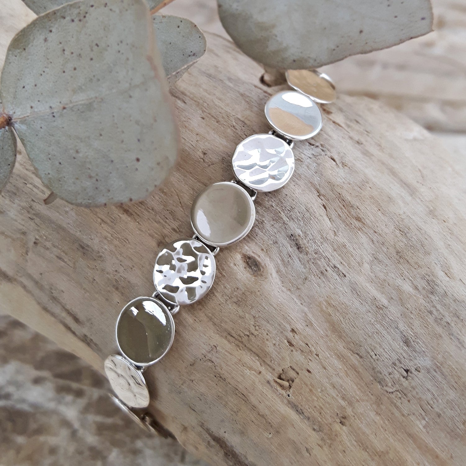 Silver bracelet with round, textured discs on a wooden surface with eucalyptus leaves.