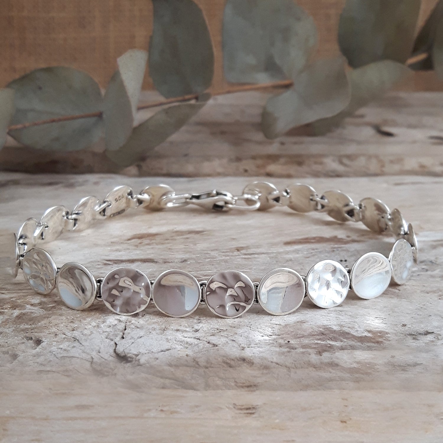 Silver bracelet with circular links on a wooden surface with eucalyptus leaves in the background
