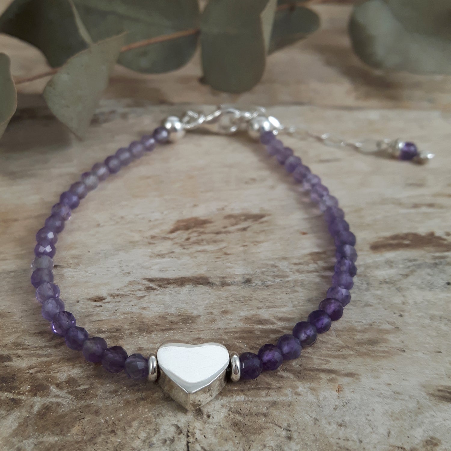 Amethyst beaded bracelet with a silver heart charm on a wooden surface with green leaves in the background