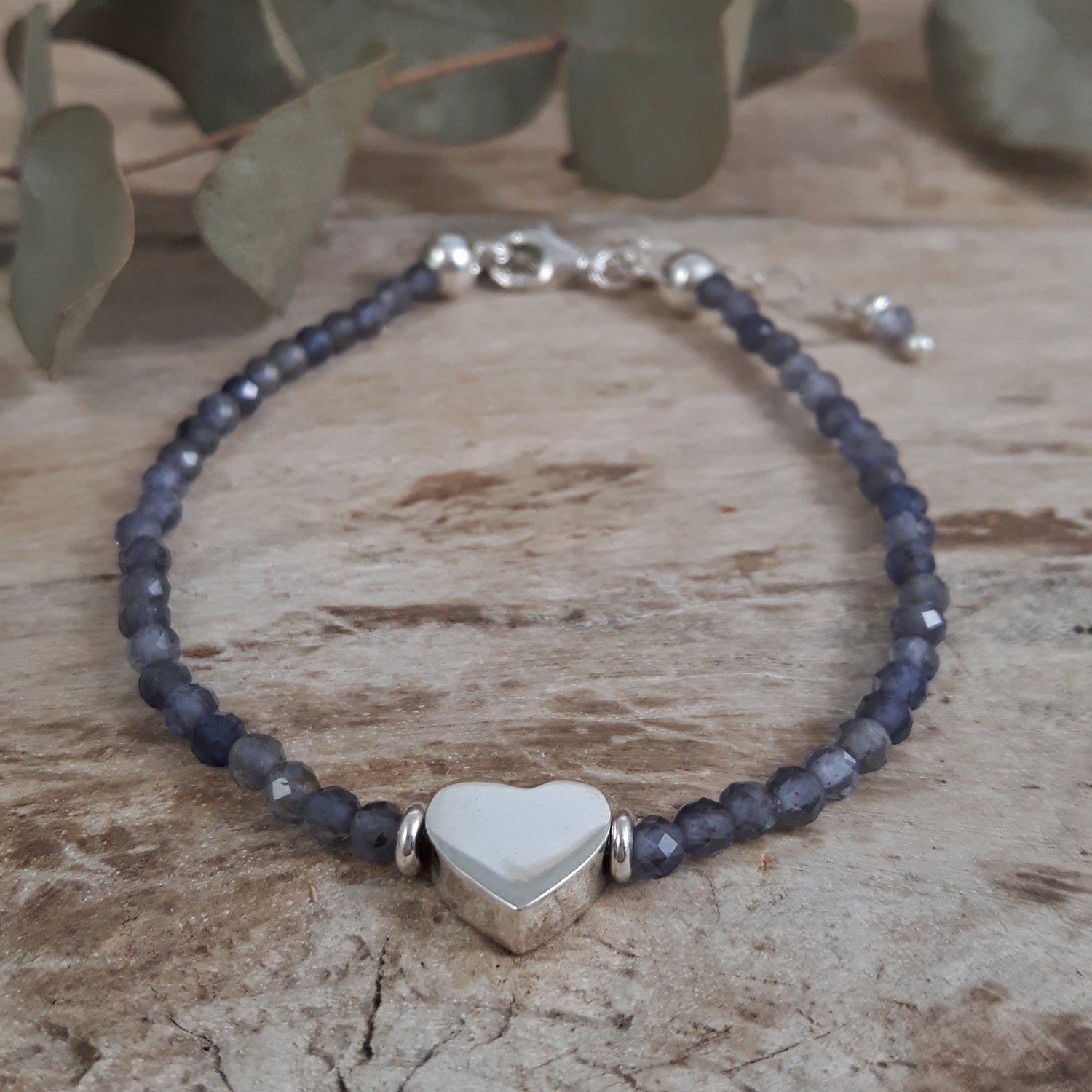 Iolite Gemstone Birthstone Bracelet with heart-shaped charm on a wooden surface with green leaves in the background