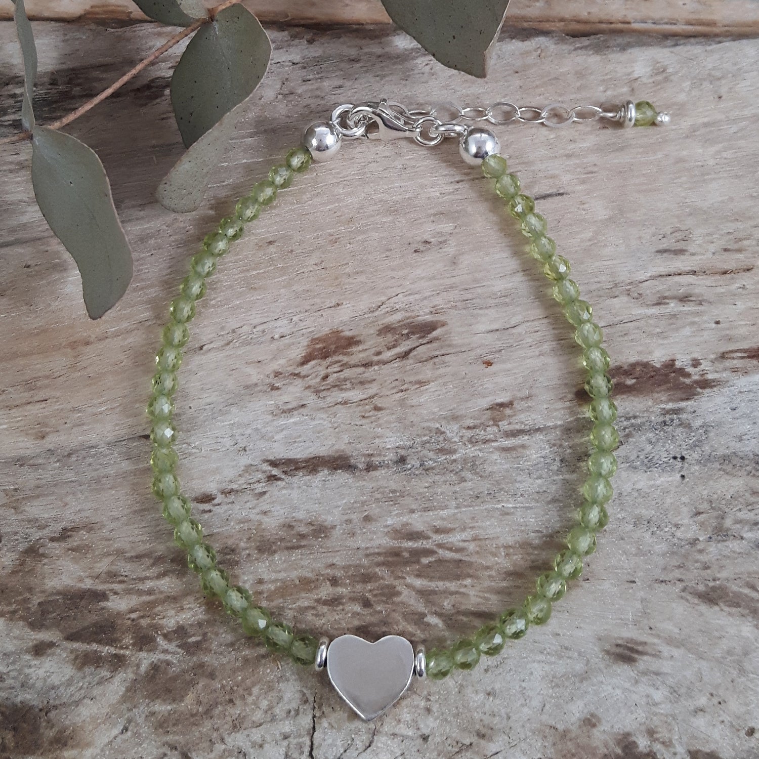 Green peridot beaded birthstone bracelet with a sterling silver heart charm on a wooden surface