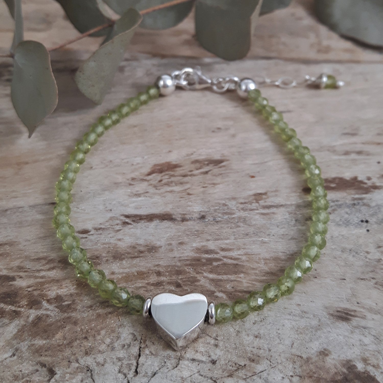 Peridot beaded birthstone bracelet with a sterling silver heart charm on a wooden surface.