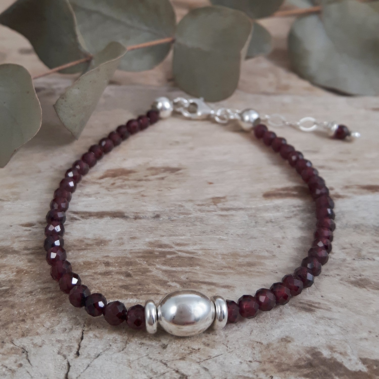 Garnet beaded bracelet with silver clasp on a wooden surface with eucalyptus leaves.