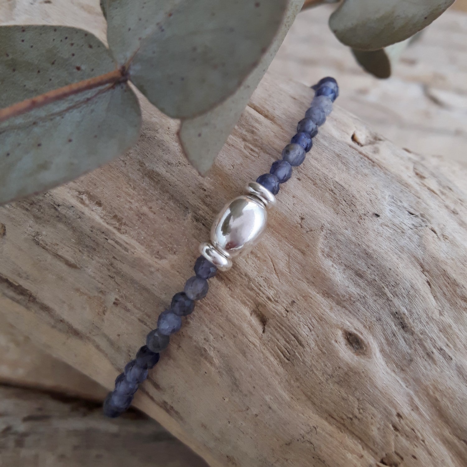 Iolite birthstone beaded bracelet with a sterling silver bead on a wooden surface with eucalyptus leaves.