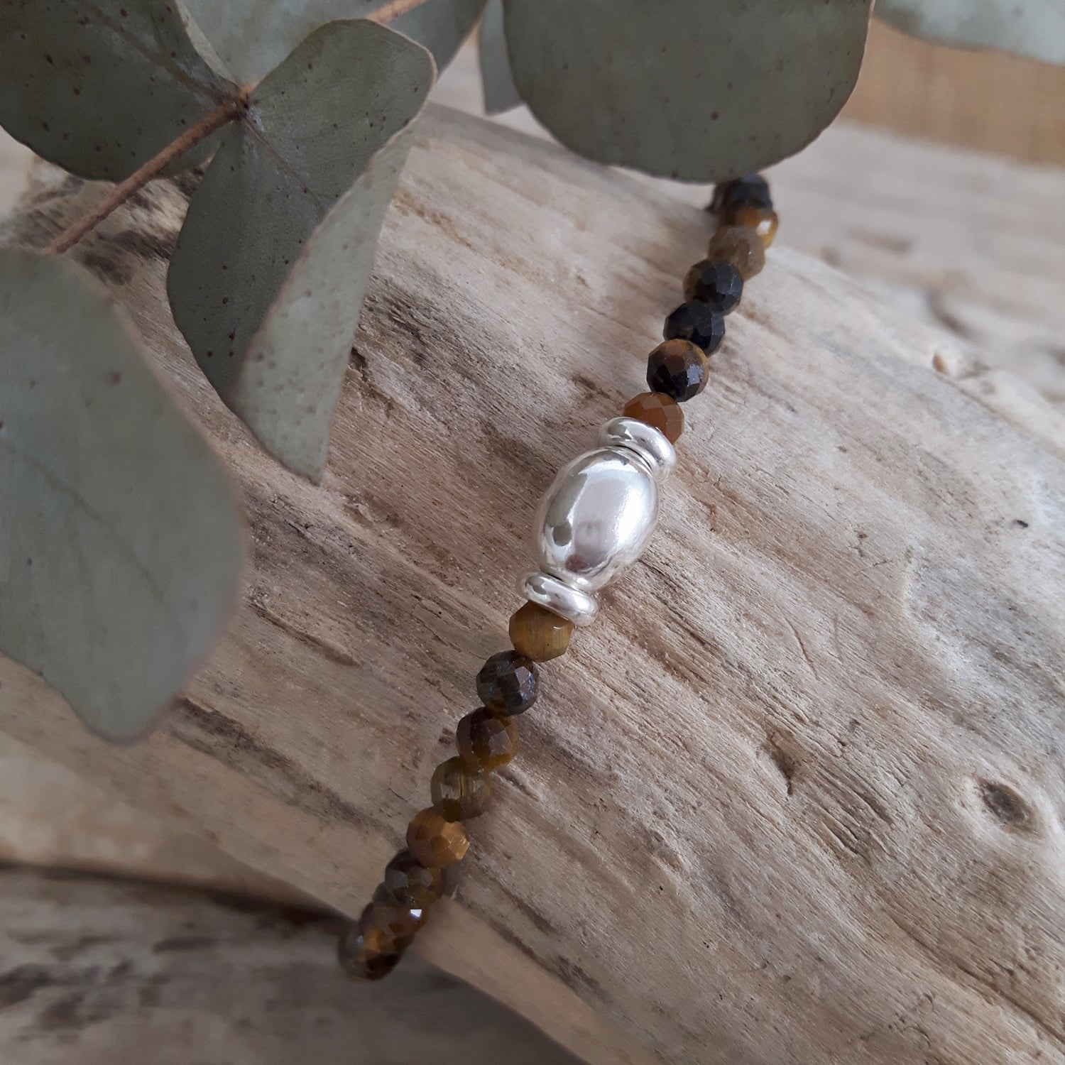 Tiger's eye beaded birthstone bracelet with a sterling silver orb bead on a wooden surface with eucalyptus leaves.