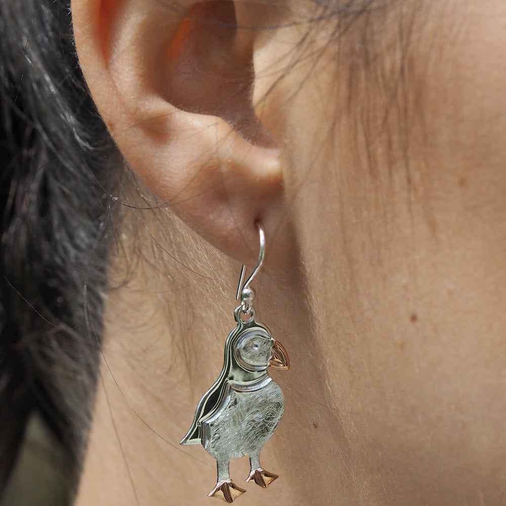 A drop-style earrings featuring a puffin design, with a mix of polished and satin finishes, predominantly silver with copper details for the beak and feet, is worn by a person.
