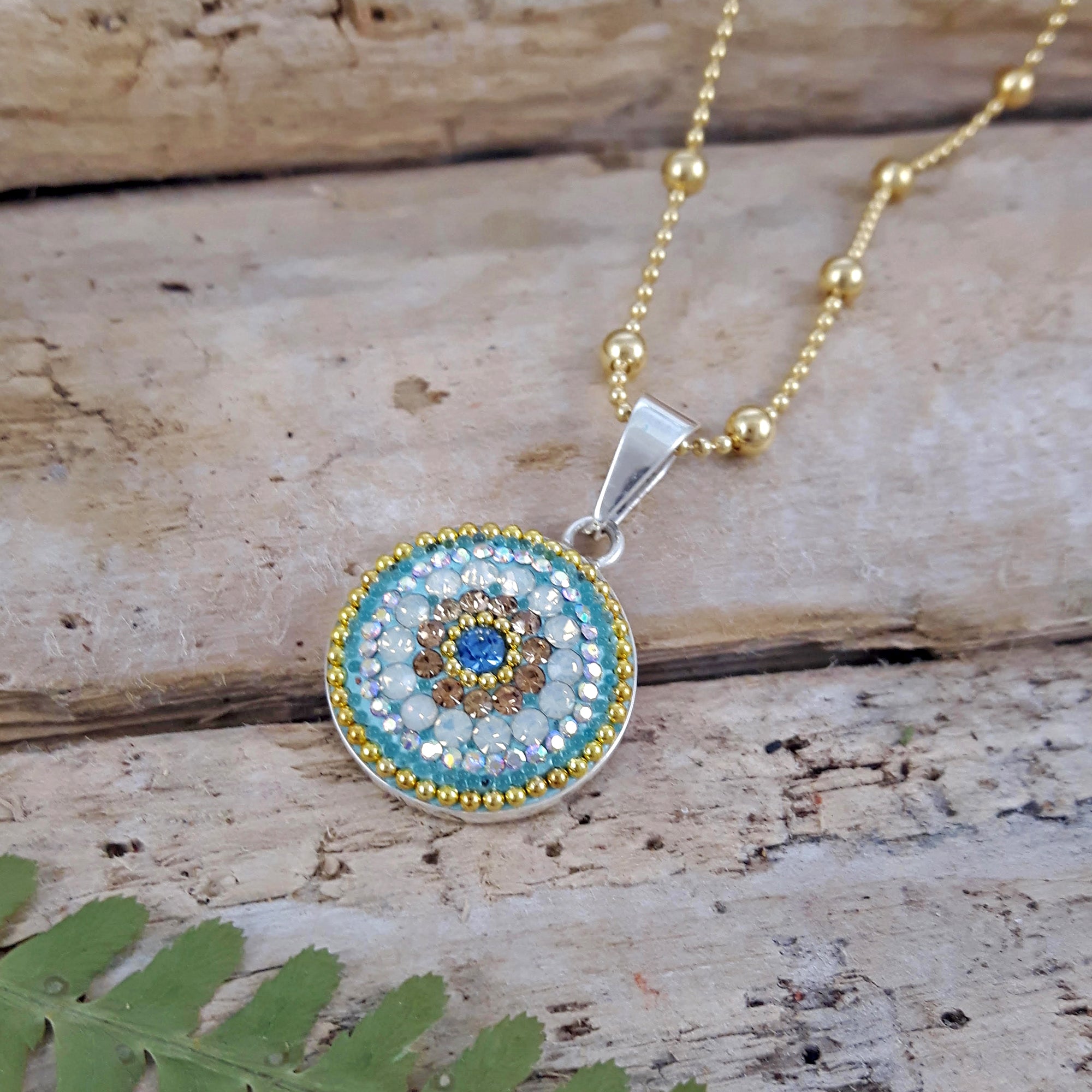 A sterling silver coin-shaped pendant with a floral pattern, featuring a mix of clear, turquoise and aqua crystals, with gold microbead detail, displayed on a gold chain.