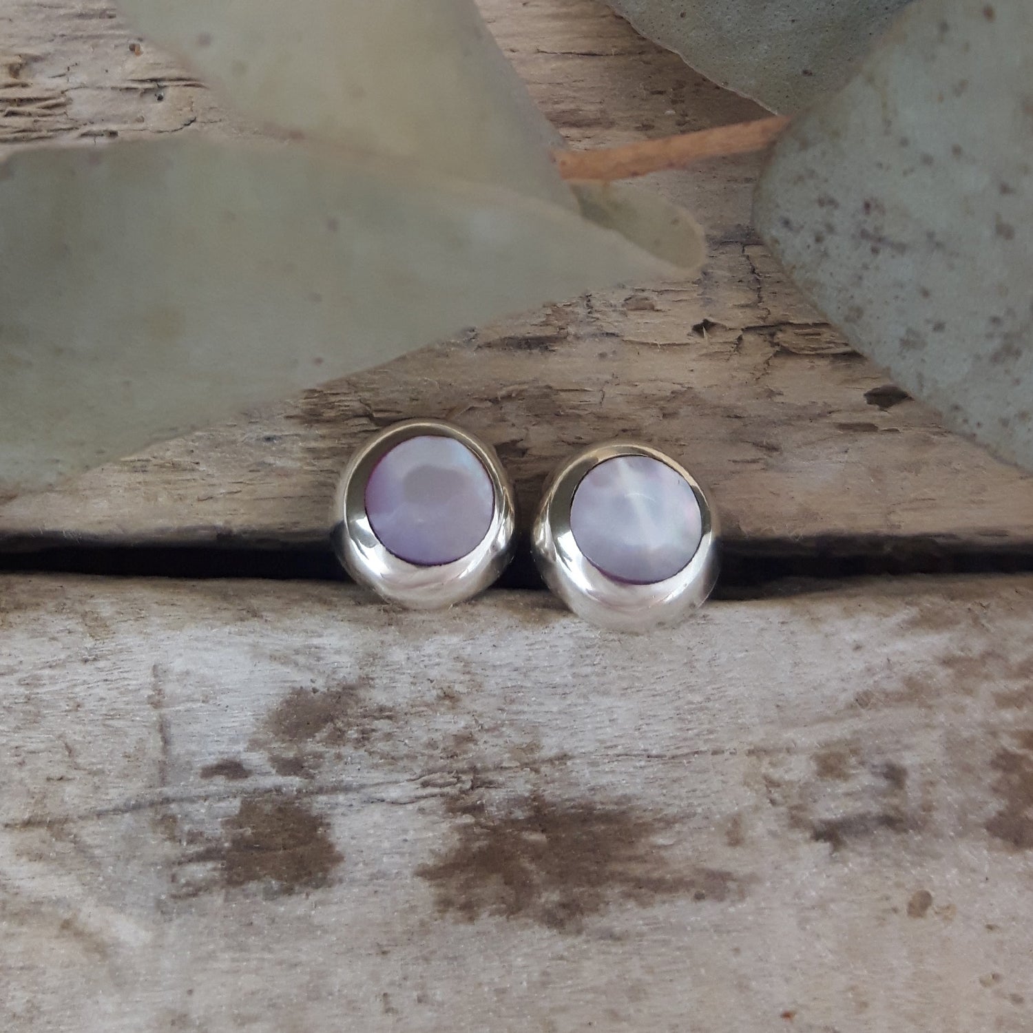 Pair of dainty pink mother-of-pearl circle stud earrings in 925 sterling silver, styled between driftwood.