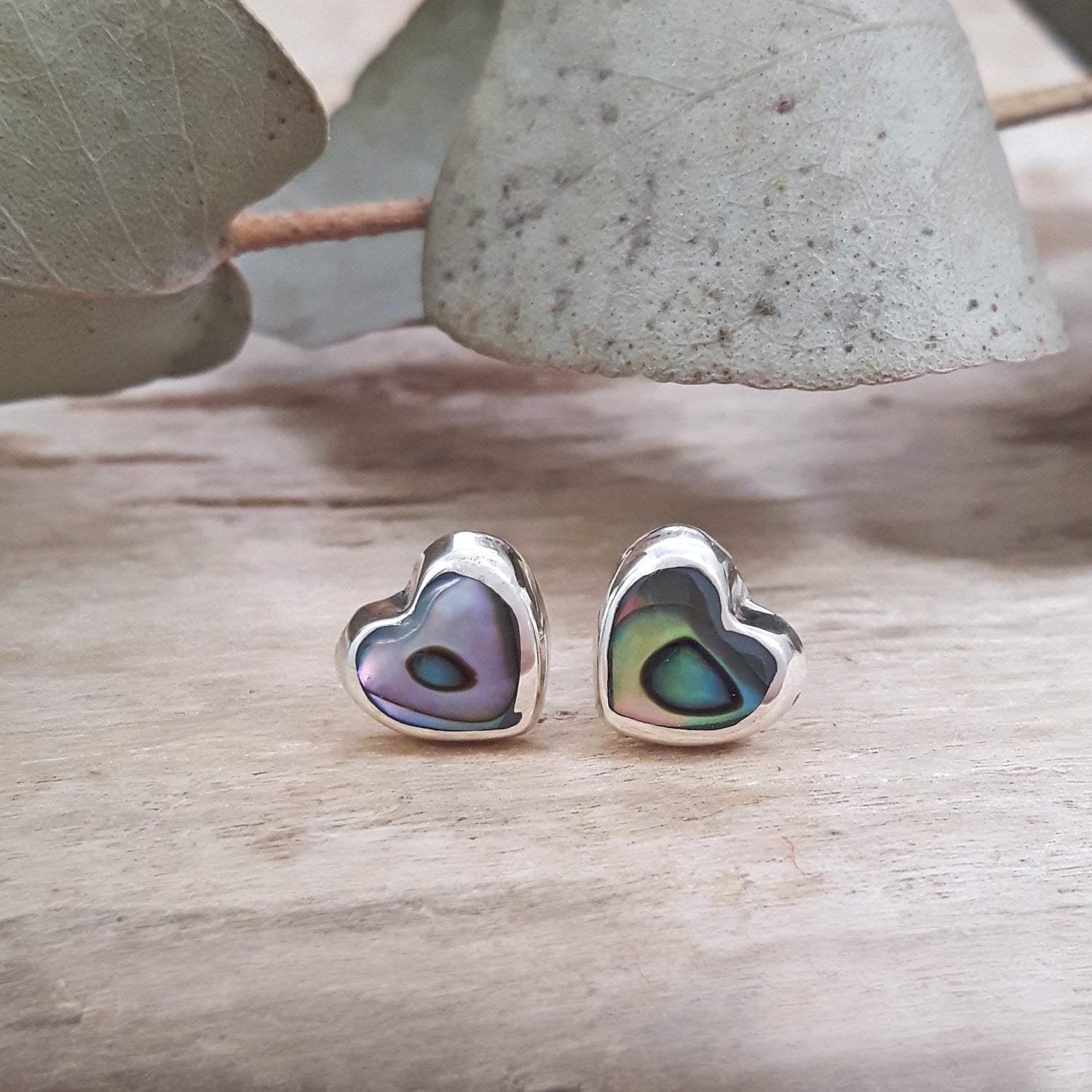 Dainty abalone heart stud earrings in 925 sterling silver, showing iridescent purple and green shell, flat-lay on wood