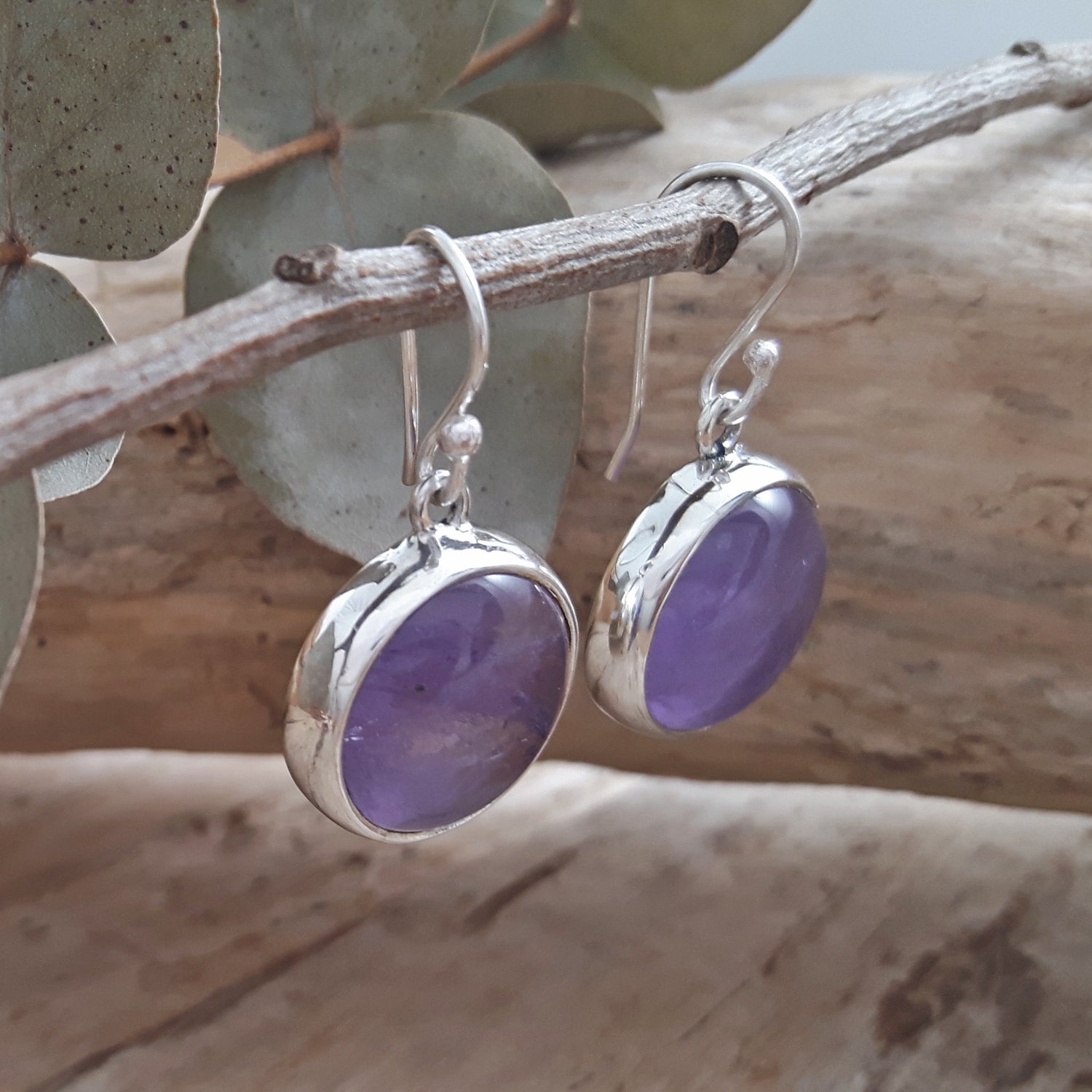 Purple amethyst gemstone earrings with sterling silver hooks hanging on a branch with leaves in the background