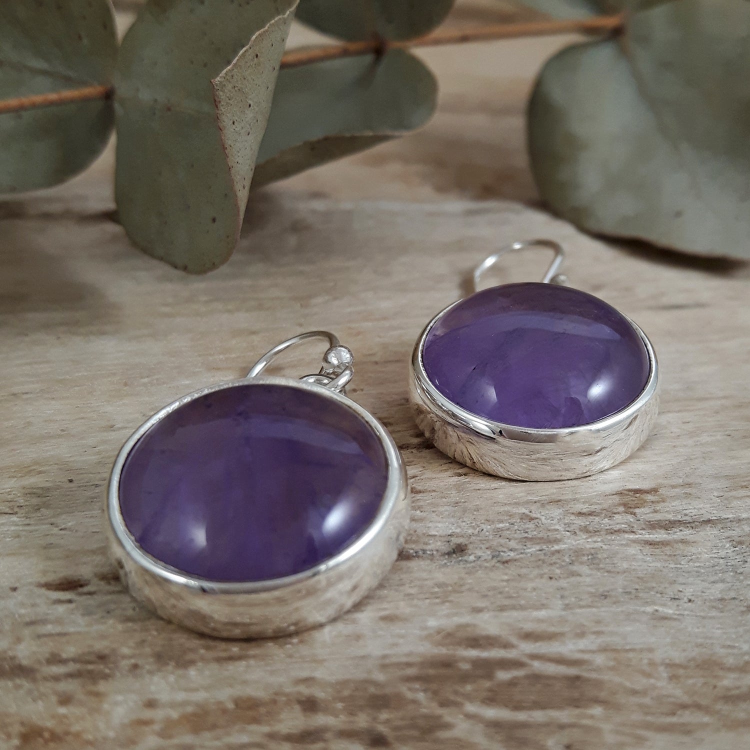 One-of-a-kind purple amethyst gemstone earrings with sterling silver rims on a wooden surface with green leaves in the background
