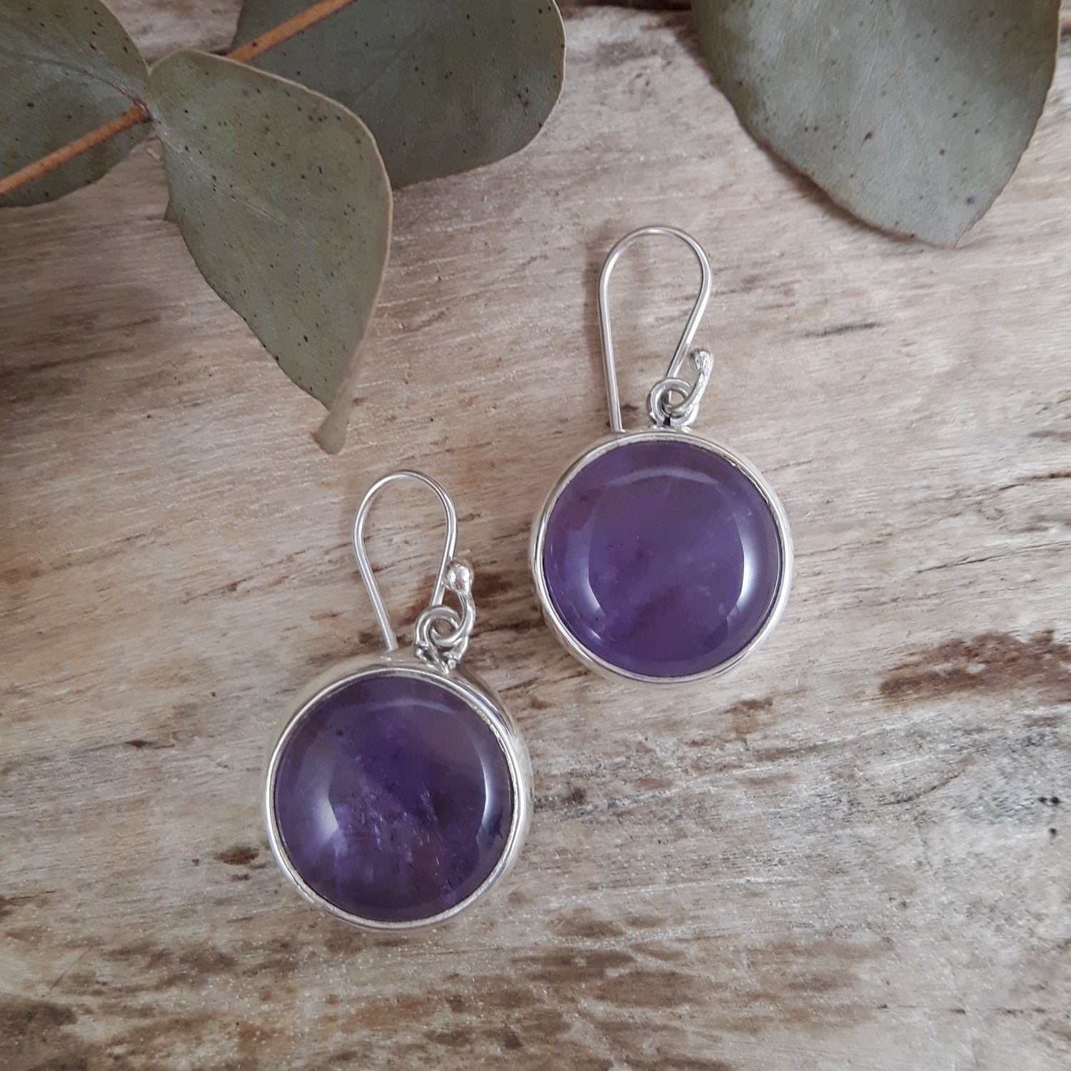 Purple round amethyst drop earrings with sterling silver rims on a wooden surface with eucalyptus leaves.