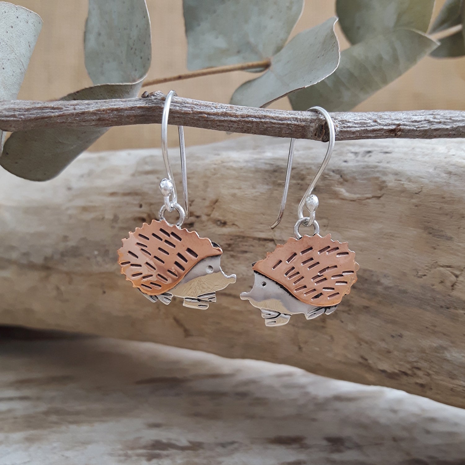 Copper and silver edgehog-shaped drop earrings on a branch with natural background