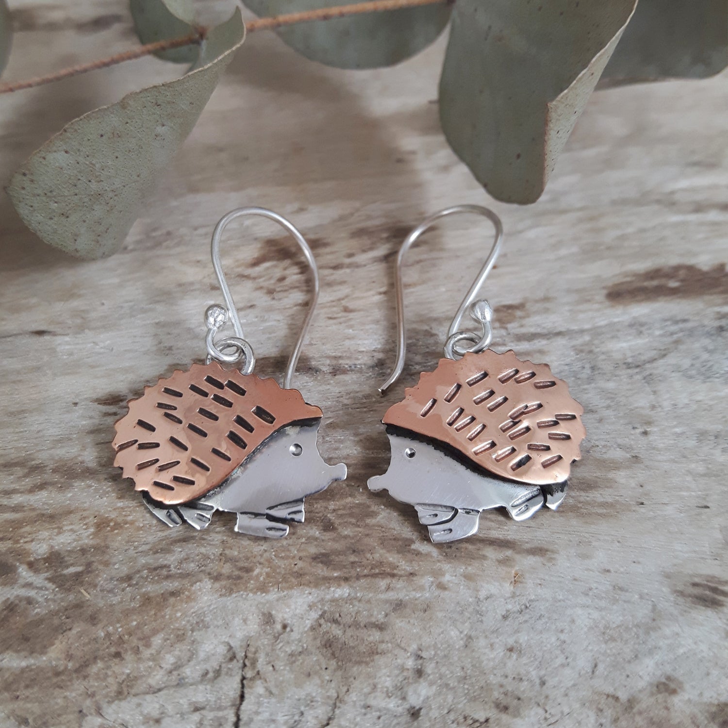 Hedgehog-shaped drop earrings with copper and sterling silver finish on a wooden surface with leaves.
