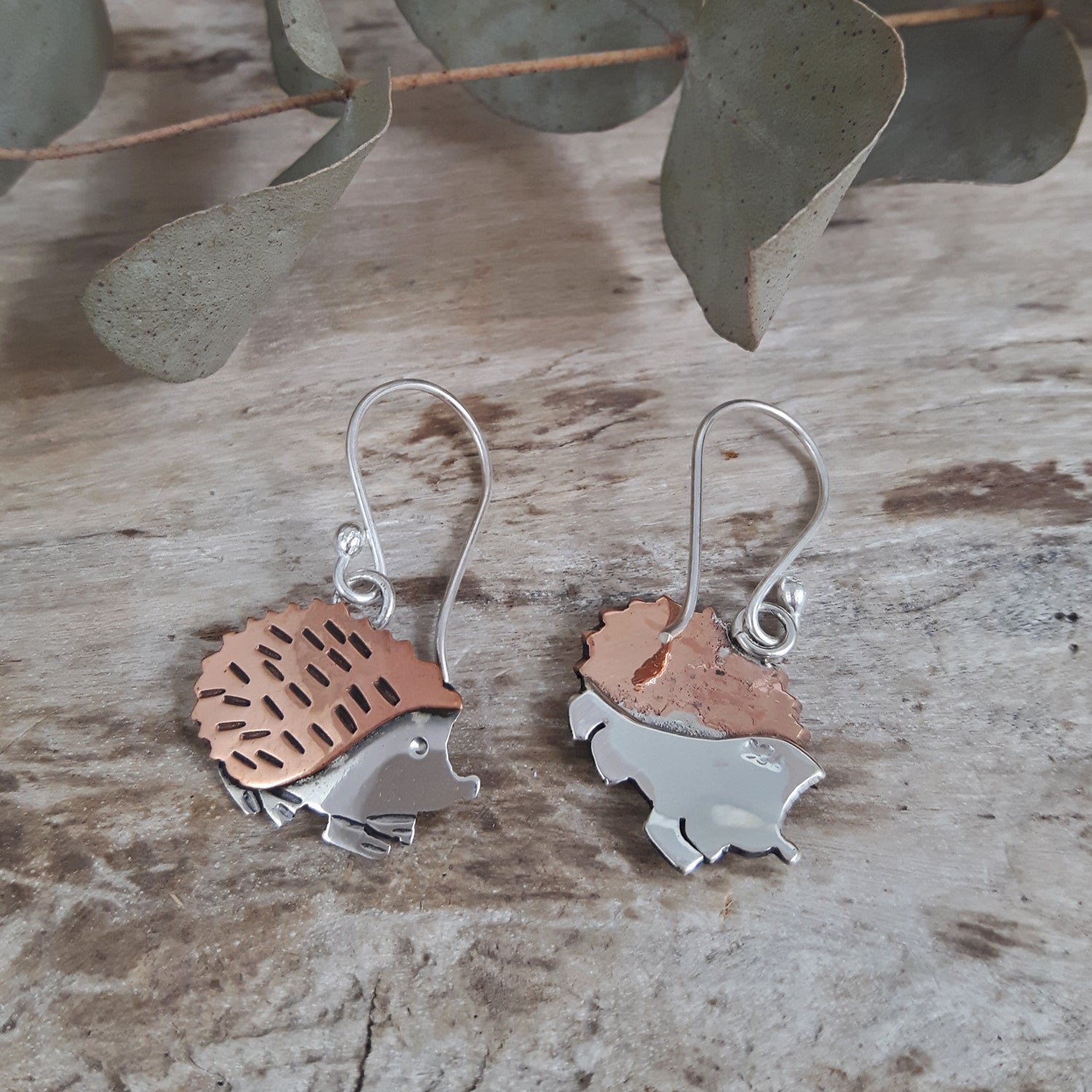 Hedgehog-shaped drop earrings with copper and sterling silver finish on a wooden surface with eucalyptus leaves.
