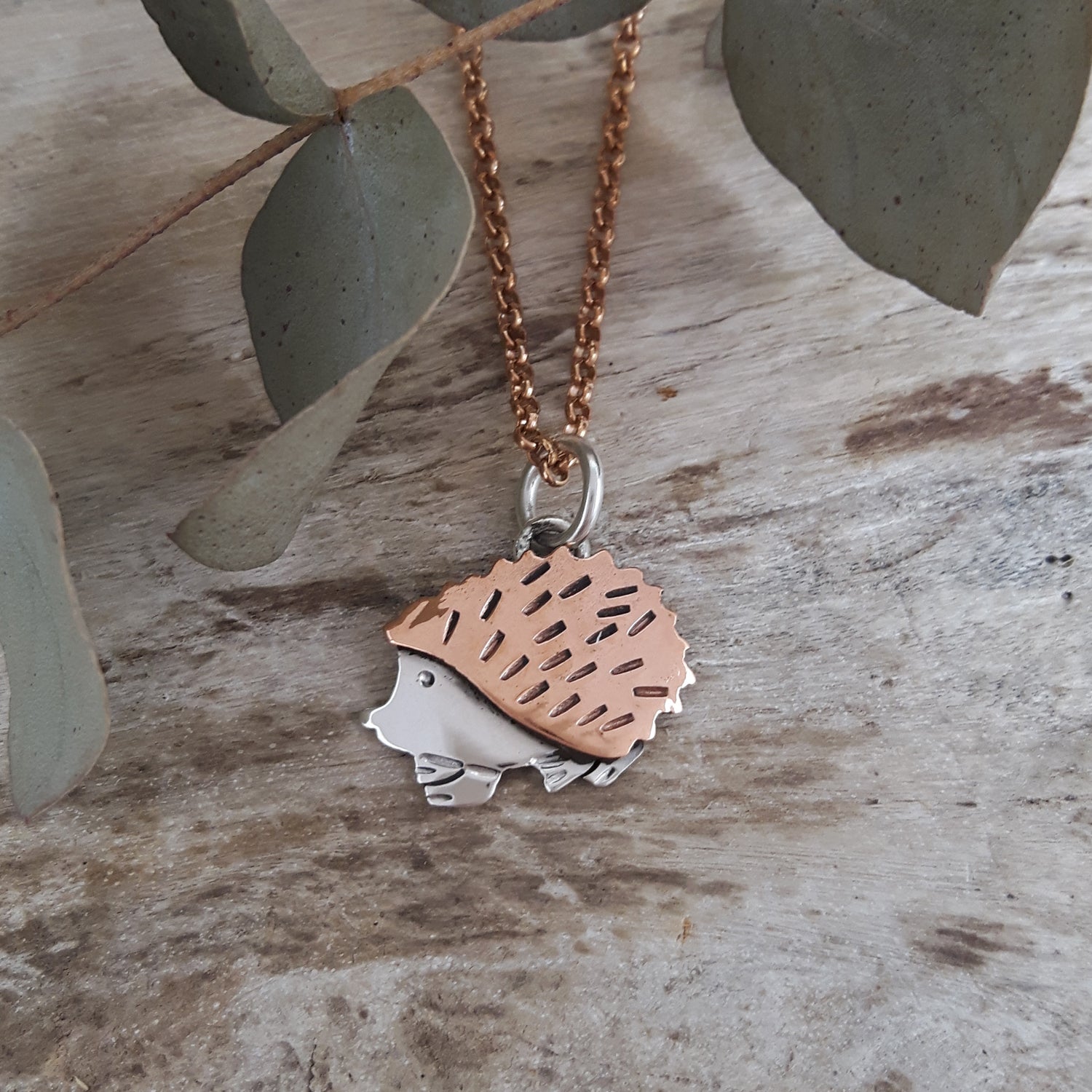 Rose gold chain with a hedgehog pendant on a wooden surface with eucalyptus leaves.