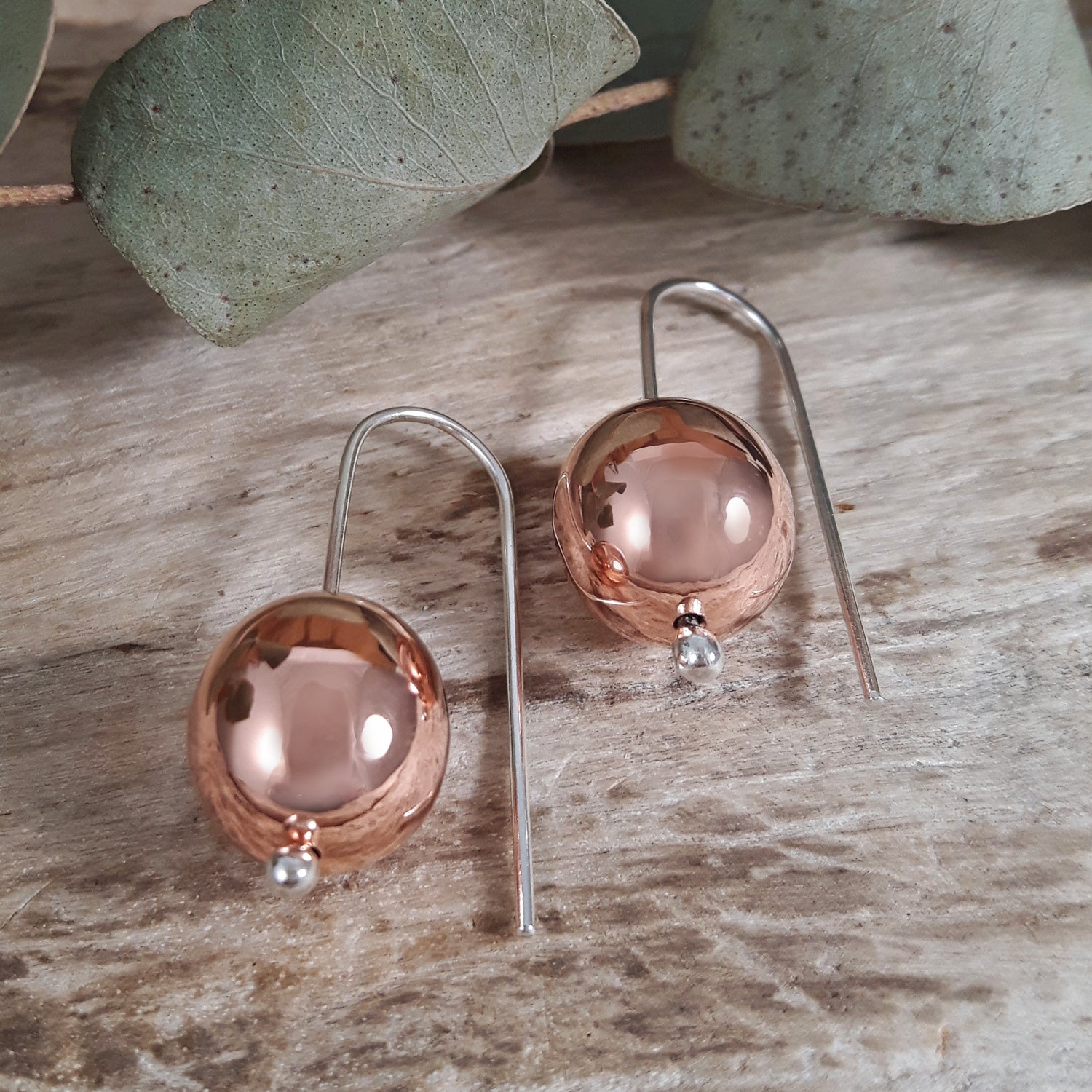 Pair of copper sphere earrings on a wooden surface with eucalyptus leaves.