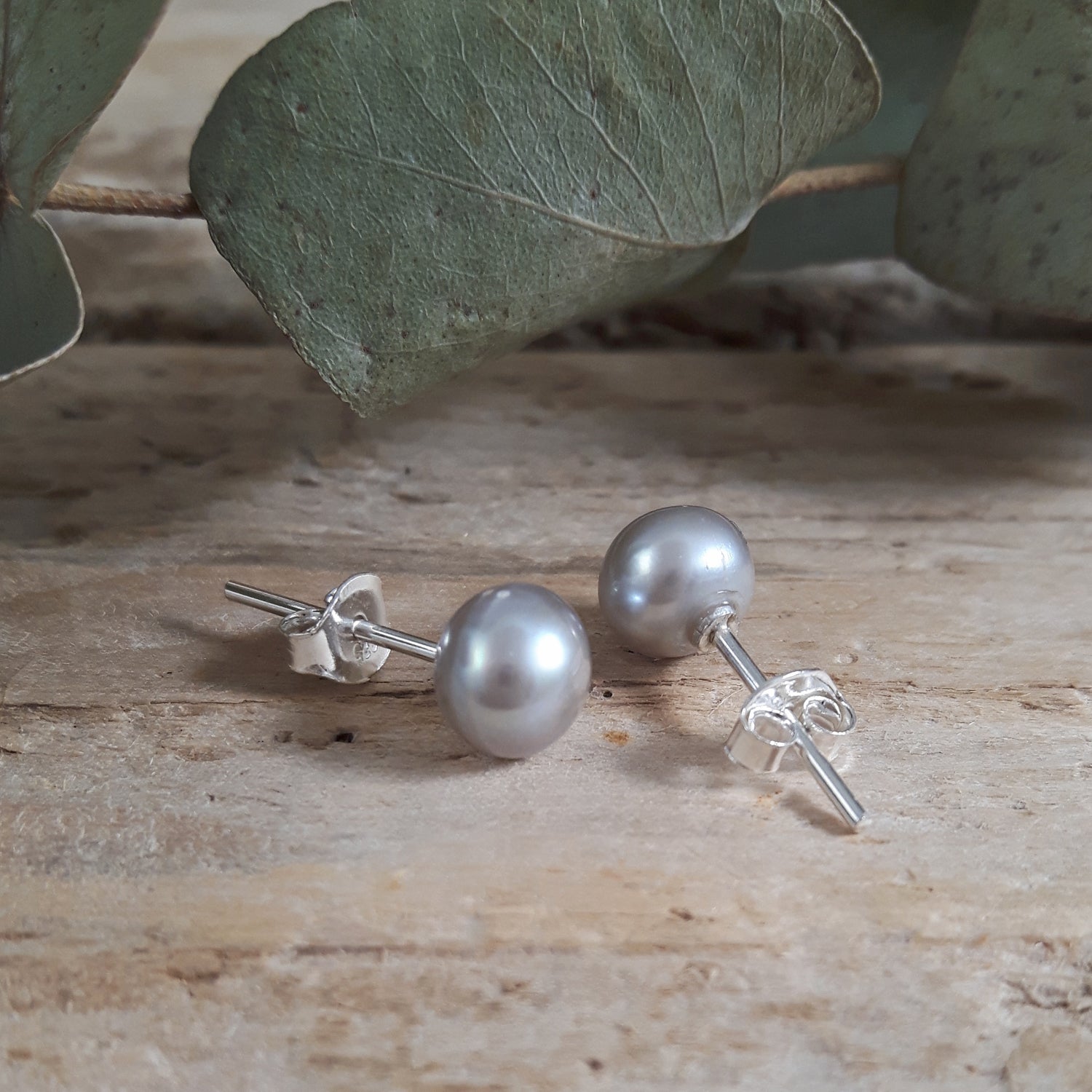 Silver pearl earrings on a wooden surface with eucalyptus leaves in the background