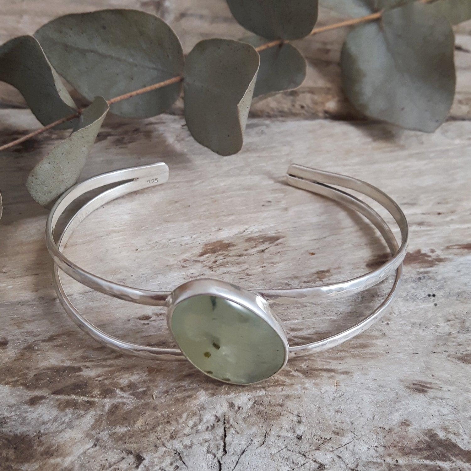 Sterling silver bracelet with a green prehnite stone on a wooden surface with eucalyptus leaves.