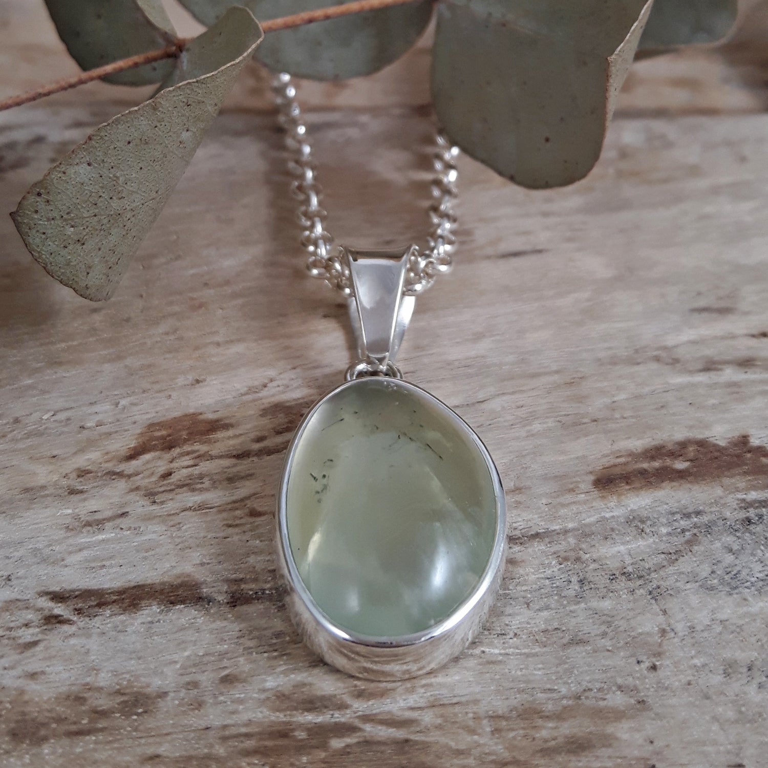 Sterling silver pendant with a green prehnite gemstone on a wooden surface with eucalyptus leaves.