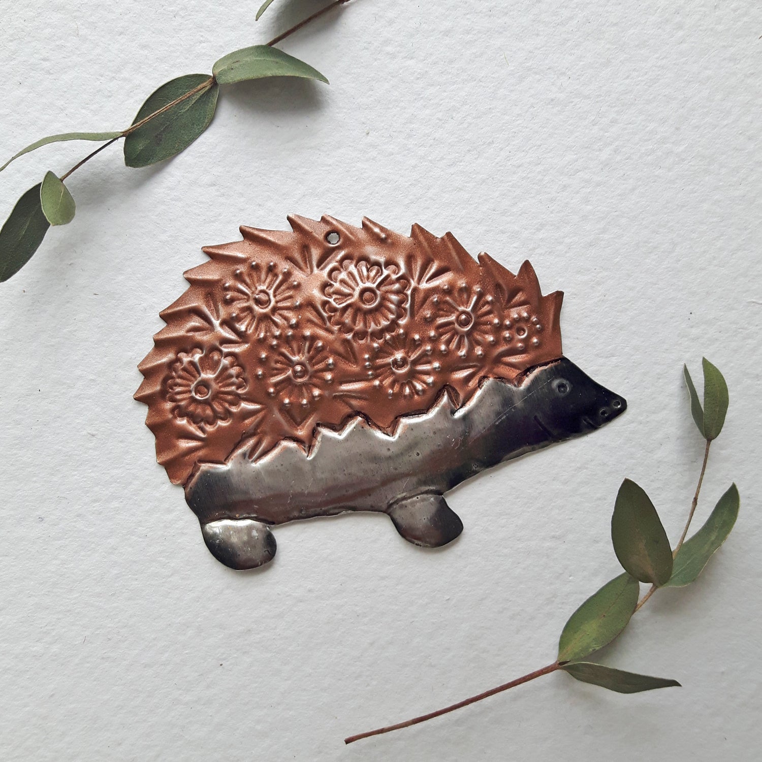 Decorative metal hedgehog ornament on a white background with green leaves.