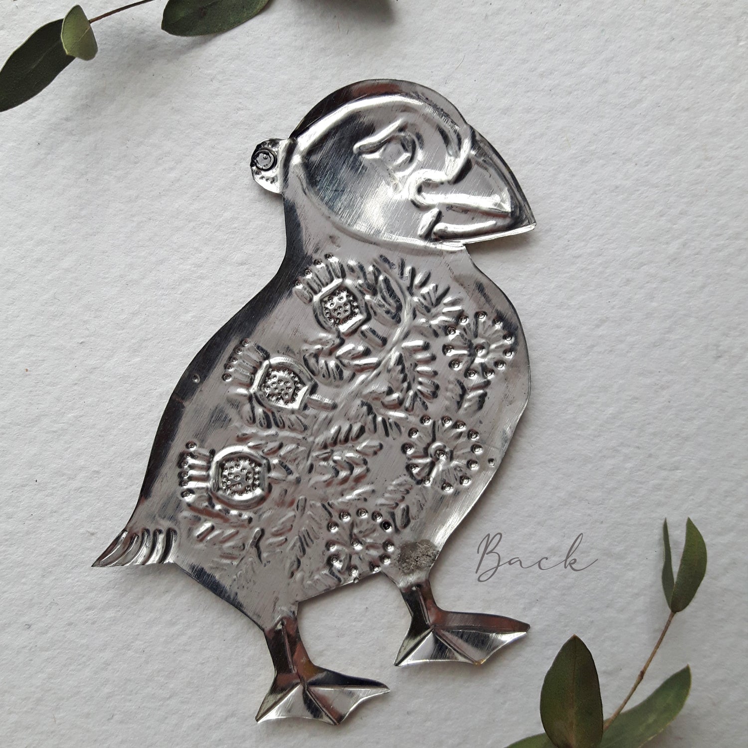 Decorative metal penguin ornament on a textured white background with green leaves.