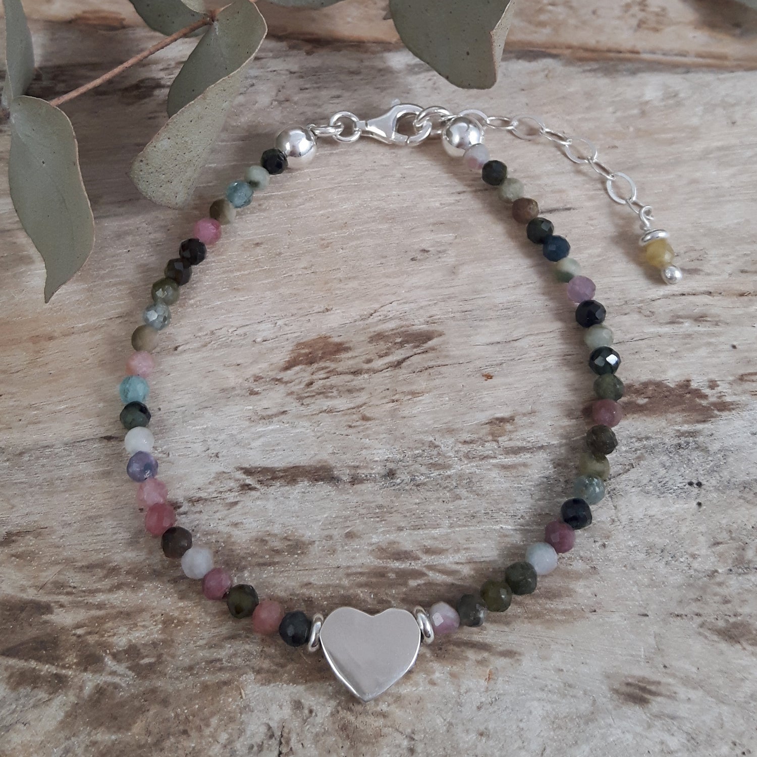 Multicoloured tourmaline birthstone beaded bracelet with a sterling silver heart charm on a wooden surface