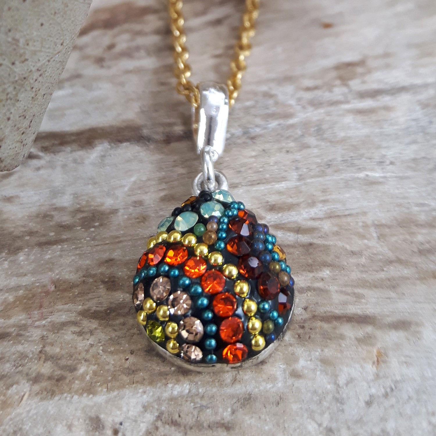 Multicoloured beaded pendant on a gold-plated chain against a wooden background. Teardrop, bohemian style Allegra California Dream pendant.